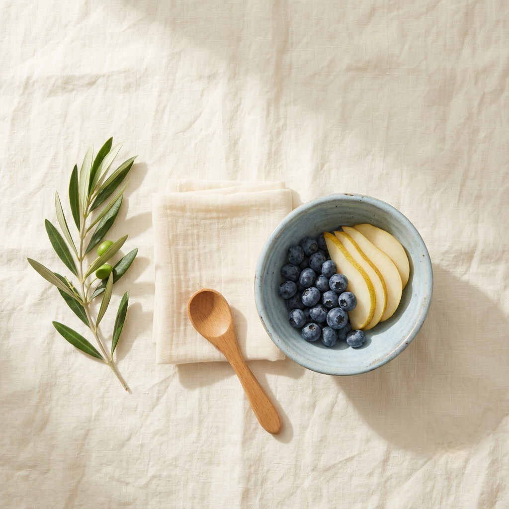 A pale blue ceramic bowl of blueberries and sliced pear beside a folded muslin cloth, a wooden toddler spoon and a sprig of olive leaves on cream linen.